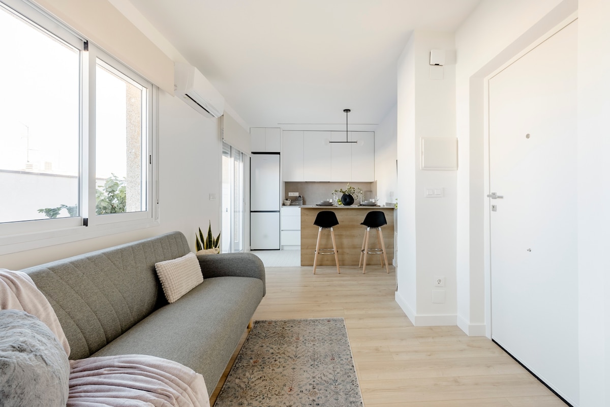 A bright living area features a comfortable gray sofa, complemented by a decorative rug. In the background, a modern kitchen is visible, equipped with sleek cabinetry and contemporary appliances. Natural light floods in through large windows, creating an open and airy atmosphere.