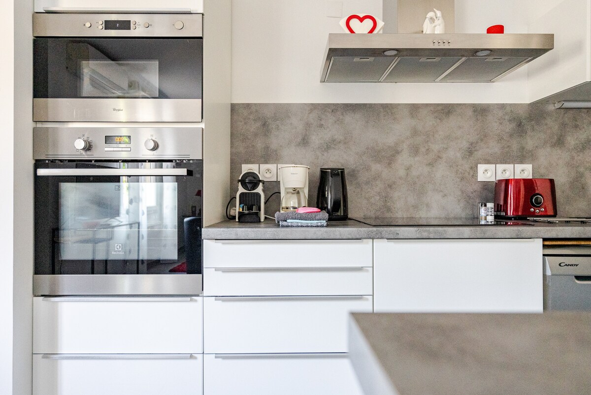 The kitchen features modern appliances, including an oven, coffee maker, and kettle, all situated against a sleek gray and white backdrop. Ample storage is provided by white cabinetry. A countertop extends for food preparation, with light reflecting off the surfaces, offering a bright and functional cooking space.