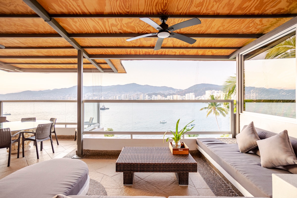 A spacious outdoor terrace overlooks the bay, featuring comfortable seating arrangements with neutral-toned cushions. A large table is positioned nearby, ideal for dining. The panorama reveals distant mountains and a calm sea, framed by swaying palm trees and large windows allowing ample natural light.