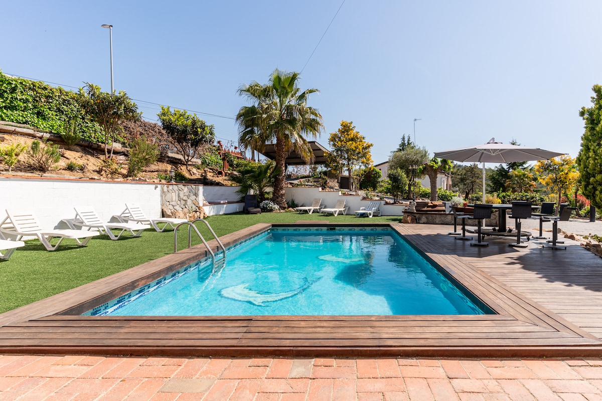 A rectangular swimming pool is framed by wooden decking, set within a landscaped garden. Sun loungers are arranged around the pool area, while a shaded seating space with an umbrella and a barbecue grill is visible in the background, creating a serene outdoor environment.