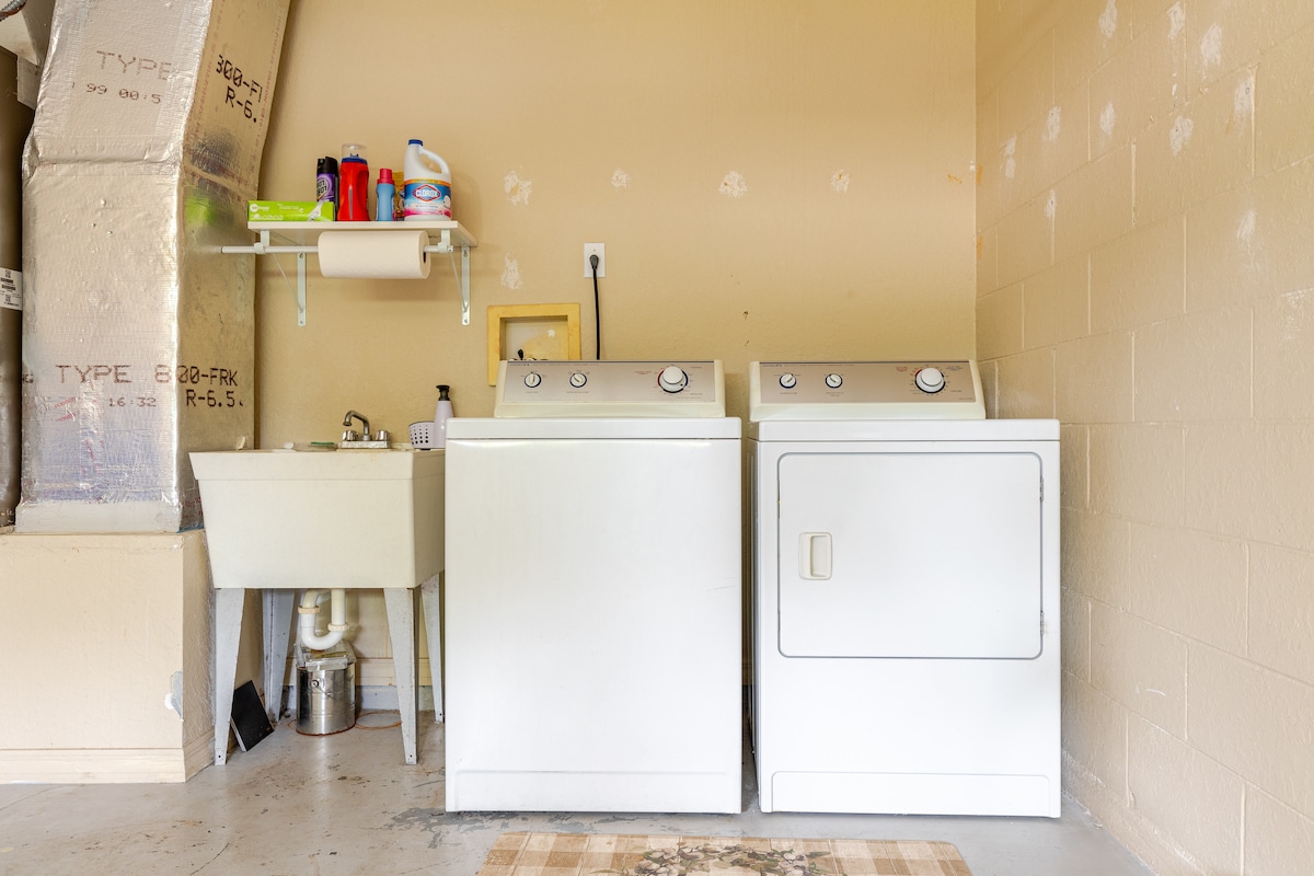 The laundry area features a washing machine and a dryer positioned side by side, with a utility sink located nearby. A shelf above holds various cleaning supplies, while a mat is placed on the floor, adding functionality to the space.