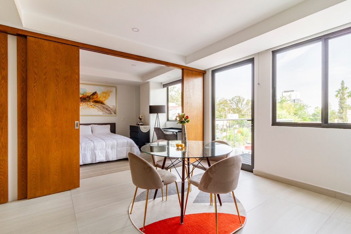 A modern living space features a round dining table surrounded by two stylish chairs. Light enters through large windows, illuminating the room. In the background, sliding wooden doors reveal a spacious sleeping area adorned with soft bedding and contemporary art.