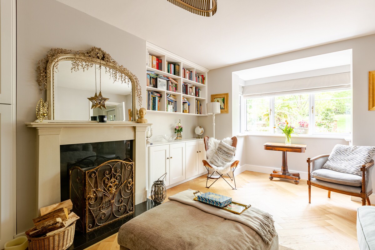 A bright living area is highlighted by natural light from a large window. A decorative fireplace with a wrought-iron screen is complemented by bookshelves stocked with various books. Cozy seating options include a butterfly chair and a small table, creating an inviting atmosphere.