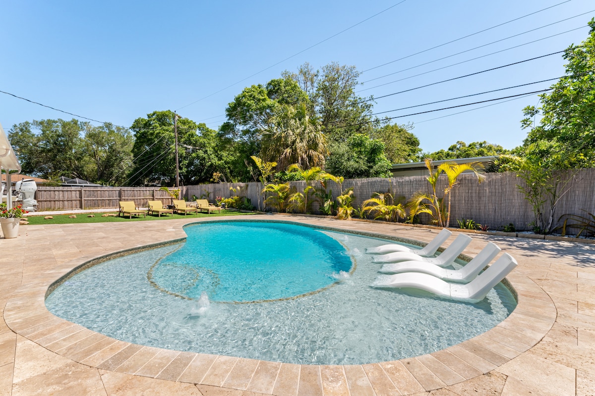 A tranquil pool area is showcased, featuring a uniquely shaped pool with a sun shelf and three white loungers. Lush greenery surrounds the space, creating a serene atmosphere. Several lounge chairs are positioned near the pool, inviting relaxation under clear blue skies.