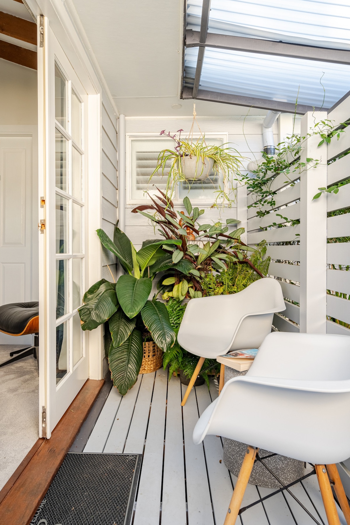 A private outdoor space is revealed, showcasing minimalist white chairs positioned on a wooden deck. Lush green plants surround the area, creating a serene ambiance. Bright light filters through the overhead canopy, enhancing the feeling of openness while ensuring a comfortable retreat.