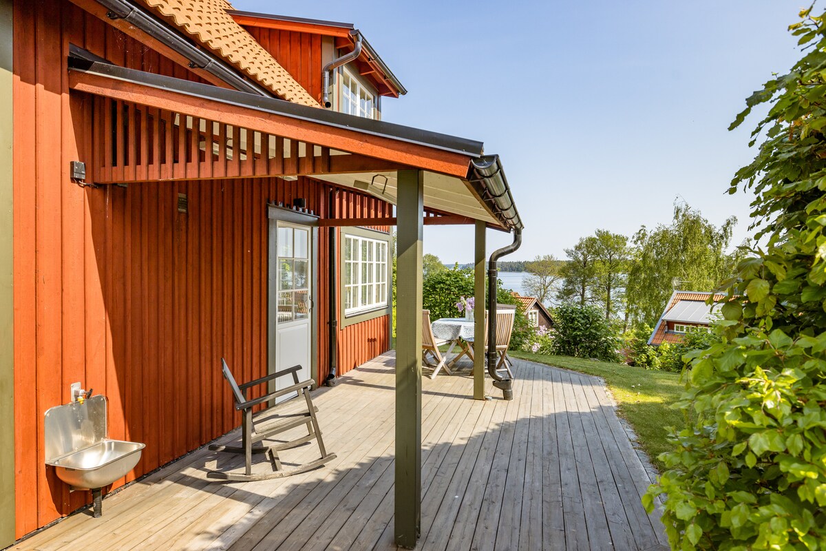 A wooden deck extends from the house, featuring a table set for outdoor dining and two chairs. A railing provides support, and the space is surrounded by lush greenery. A scenic view of nearby water can be seen in the background, enhancing the tranquil setting.
