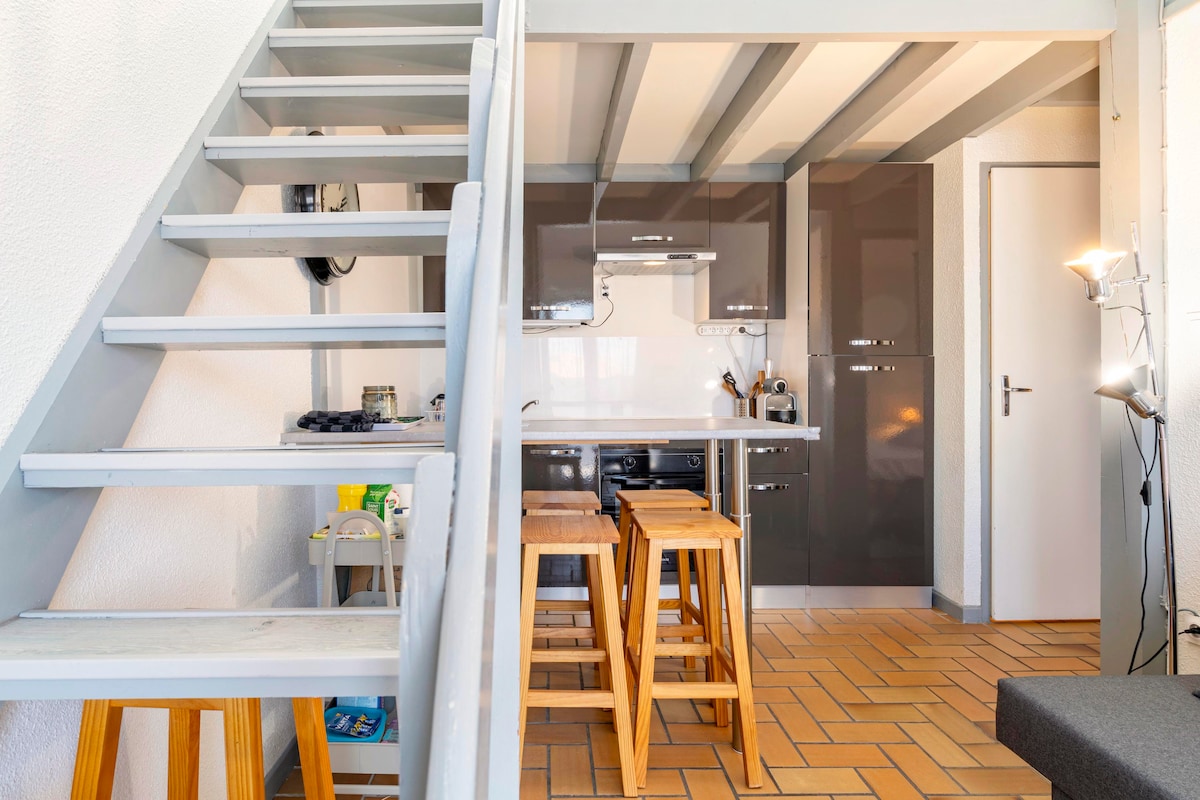 A staircase leads to the lofted area, with light grey railings contrasting against beige tiled flooring. The open kitchen features modern appliances and bar stools, while a door on the right indicates access to additional rooms. Soft lighting enhances the welcoming ambiance.