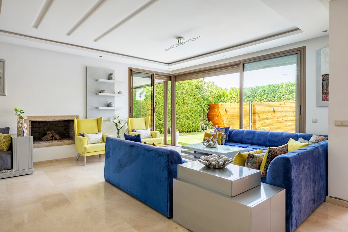 A bright living area is presented, featuring a large blue sectional sofa complemented by yellow accent chairs. Floor-to-ceiling glass doors open to a maintained garden, allowing natural light to fill the space. A modern coffee table sits at the center, accompanied by decorative elements.