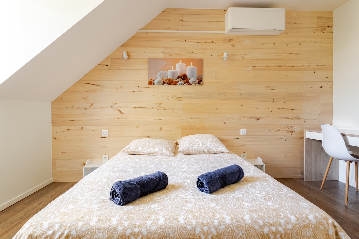A cozy bedroom features light wood paneling and a spacious bed adorned with patterned bedding. Two rolled towels are placed on the bed, and a simple desk with a chair is located against the wall, creating a calm and functional space.
