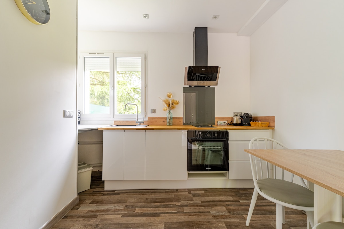 A modern kitchen features a wooden countertop and sleek white cabinetry. Natural light enters through two windows, illuminating the space. An oven and stovetop are integrated into the design. A small dining table with light-colored chairs is positioned nearby, enhancing the functionality of the area.