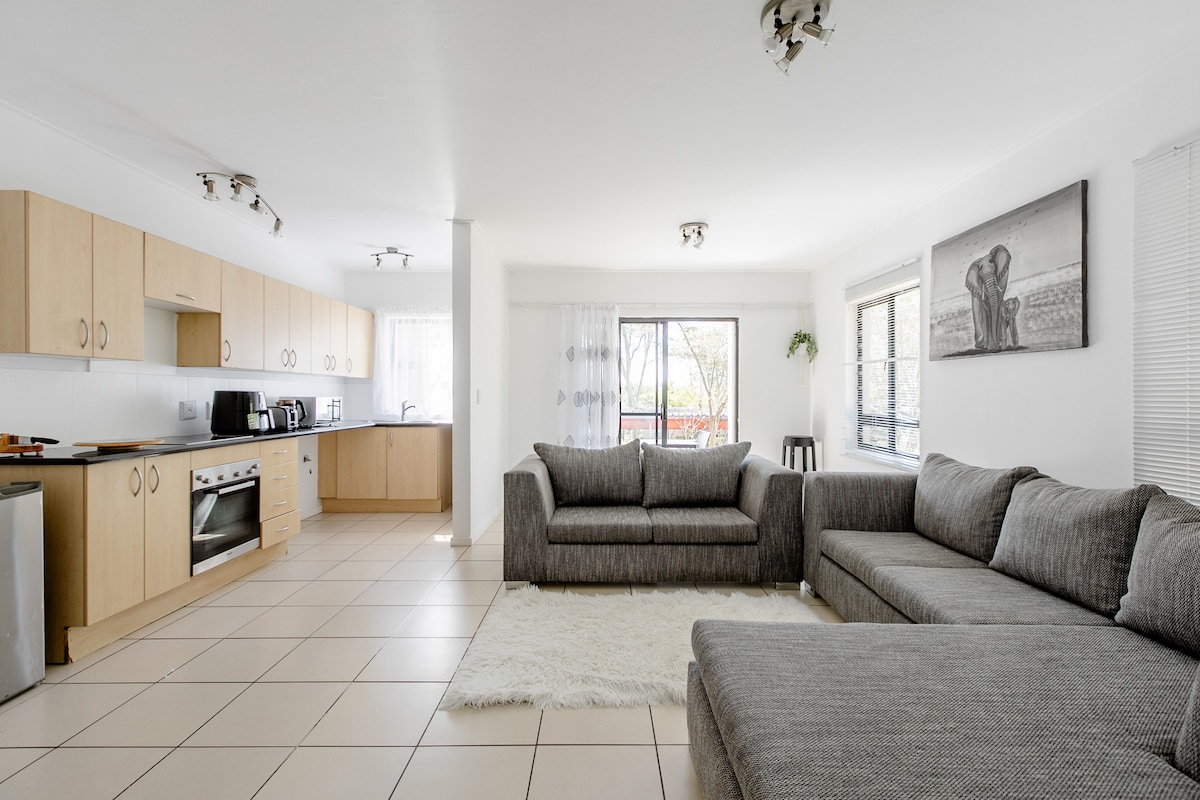 An open-plan kitchen and lounge area features light wood cabinetry and modern appliances. A spacious L-shaped couch is positioned on a soft area rug, complemented by large windows that allow natural light into the room, creating an inviting place for relaxation or dining.
