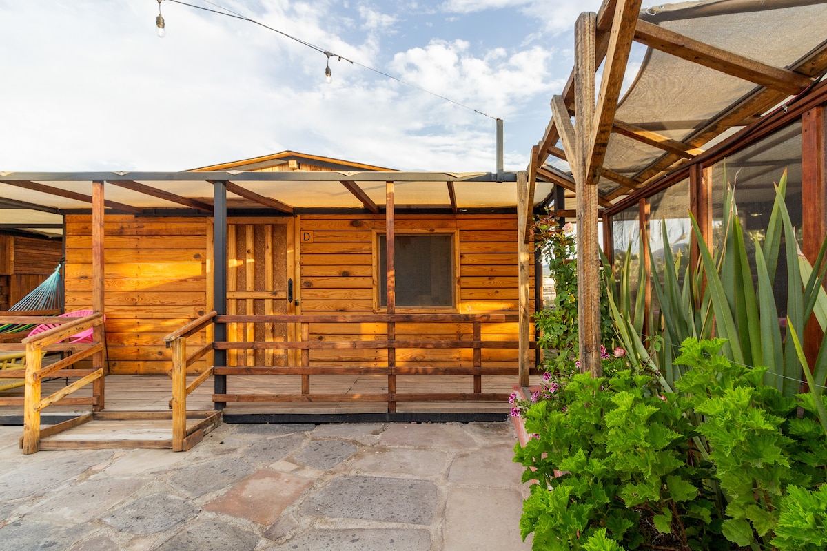 The rustic cabin exterior is constructed with natural wood, featuring a spacious porch with a railing. A hammock hangs invitingly, while vibrant green plants and flowers add a touch of color. Soft lighting creates a welcoming atmosphere as the sun sets.