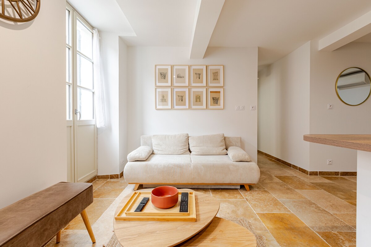 A bright living area features a light-colored sofa and a wooden coffee table with a bowl and remote control. Artwork is displayed on the wall above the couch, while a window allows natural light to flow into the space.