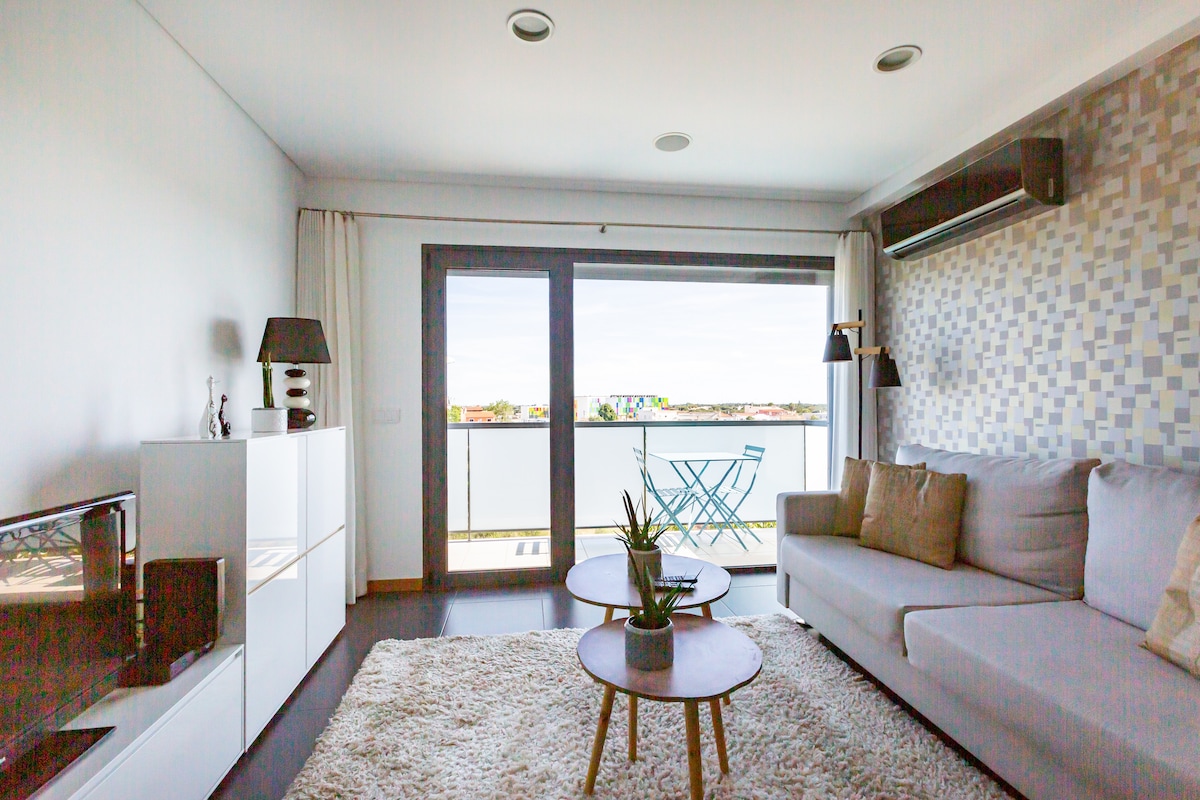 A spacious living area is furnished with a light gray sofa and two round coffee tables atop a soft area rug. Large sliding glass doors provide access to a balcony with outdoor seating, while neutral-toned decor and minimalistic accessories enhance the contemporary feel.