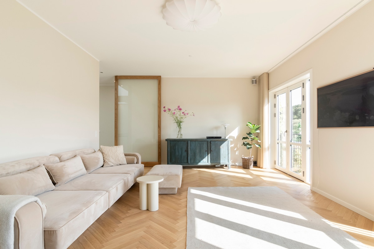 A light-filled living area features a beige sectional sofa positioned beside a minimalist white side table. A decorative cabinet stands against one wall, while a large flat-screen television is mounted above it. Natural light filters through glass doors leading outdoors.