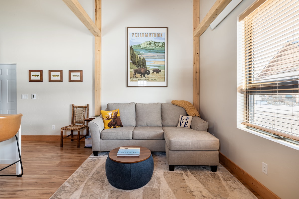 A spacious living area features a gray sectional sofa adorned with decorative pillows. A round wooden coffee table sits in front, complemented by a light area rug beneath. Natural light filters through large windows, highlighting the artwork of Yellowstone National Park above the sofa.