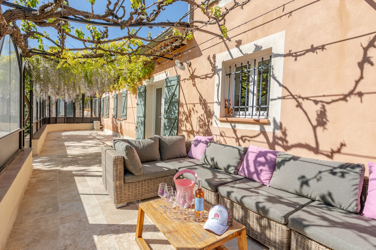 A spacious outdoor terrace is sheltered by a pergola, providing shade from the sun. Comfortable seating with soft cushions is arranged around a wooden table, accompanied by glasses and a bottle. Lush greenery frames the area, creating a relaxed environment for lounging.