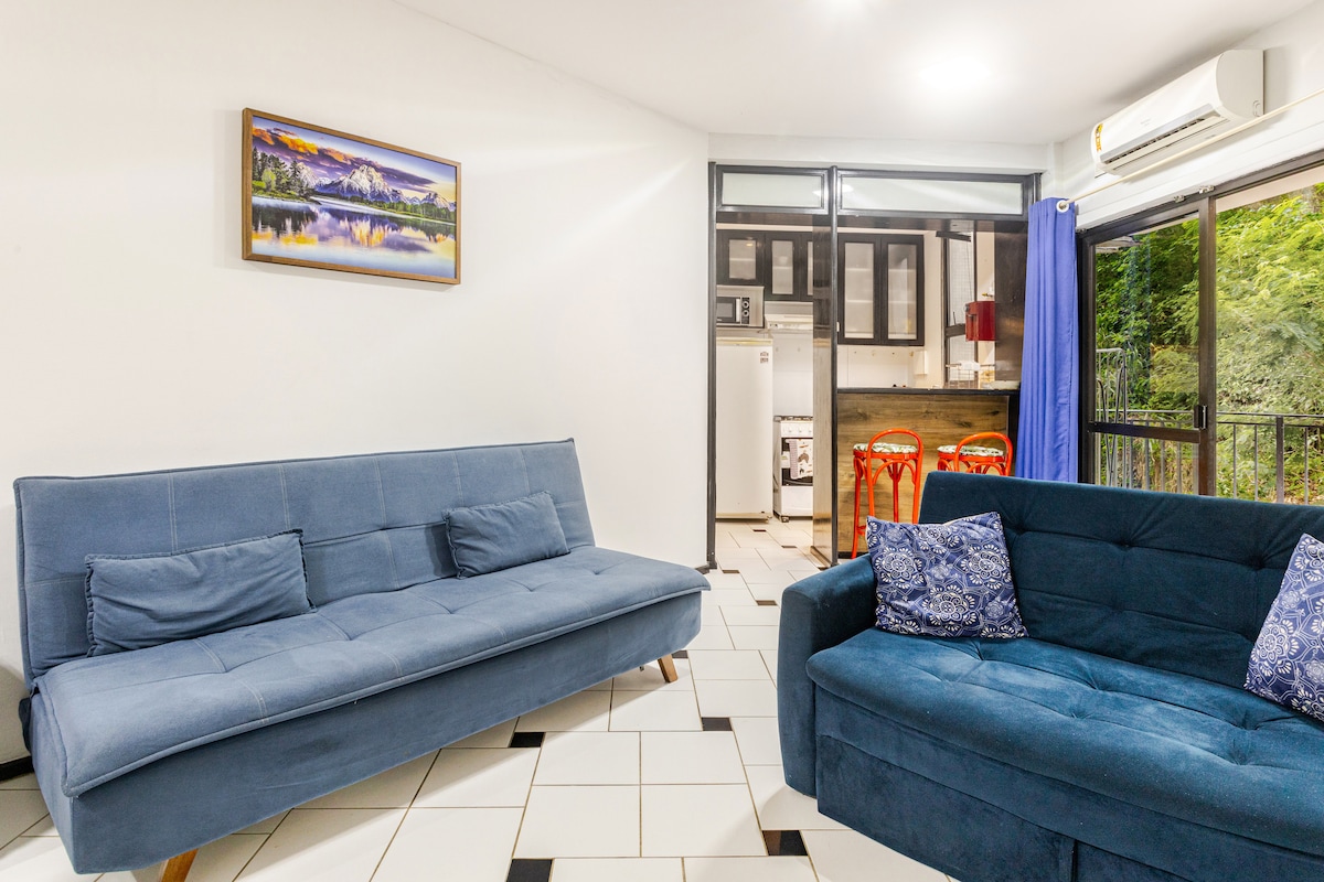 A cozy living space features two blue sofas, one of which is a convertible style. An artwork depicting a landscape is hung on the wall, and large glass doors allow natural light to fill the room while providing access to the outdoor area.