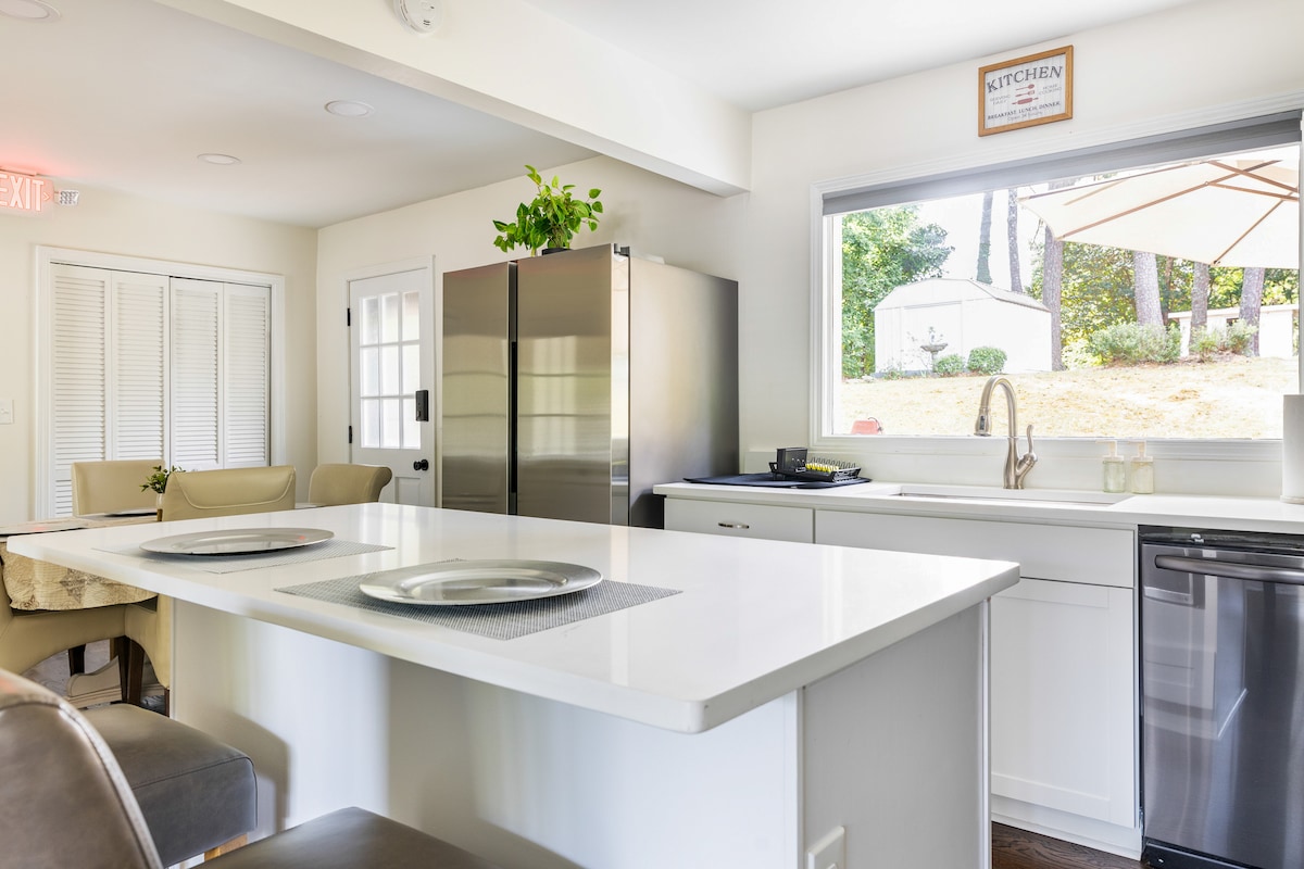 A modern kitchen is visible, featuring sleek white countertops and stainless steel appliances. A large window allows natural light to illuminate the space, where two bar stools are positioned at the island. Potted greenery adds a touch of freshness to the decor.