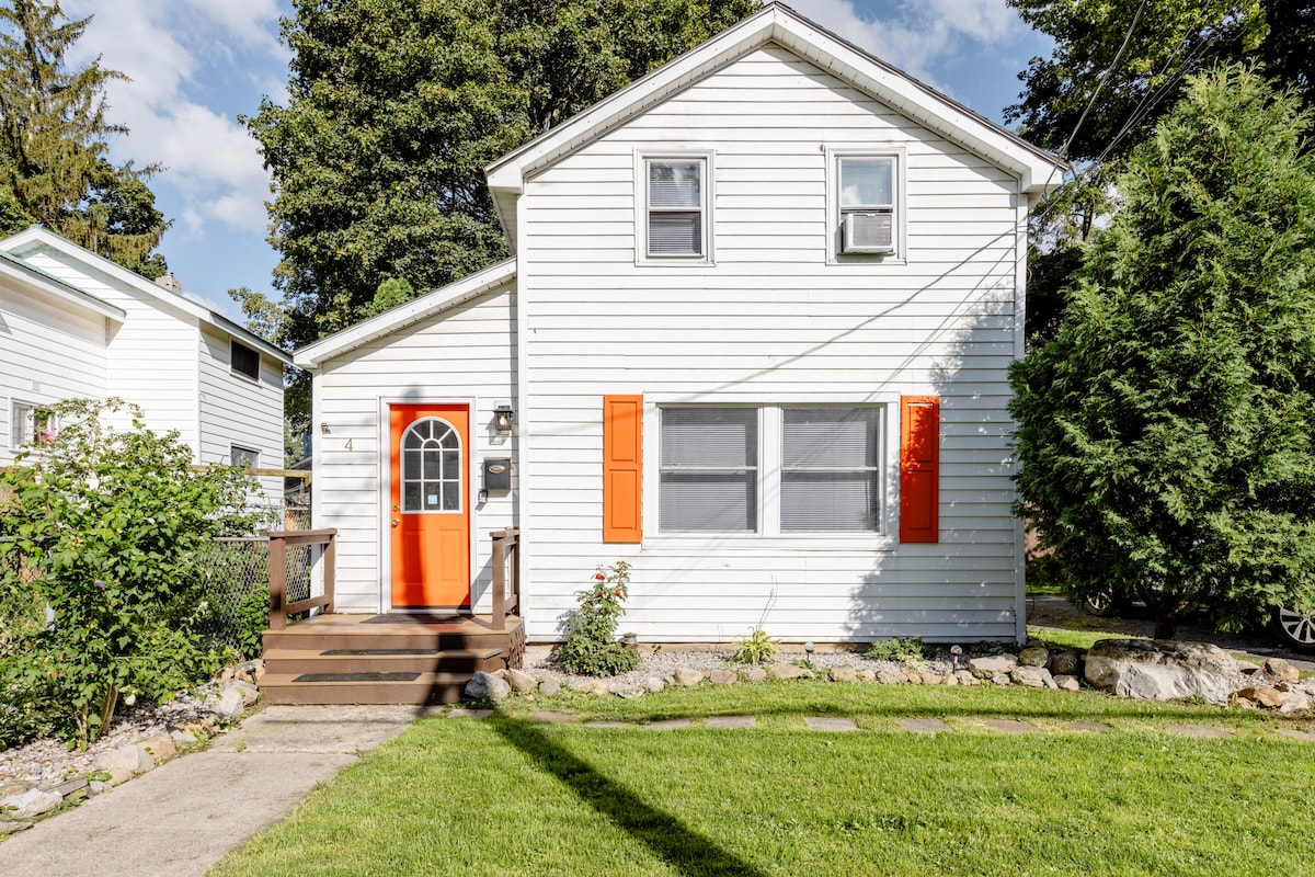 The exterior of a two-story home is depicted, featuring a bright orange front door and white siding. Lush green grass contrasts with the well-maintained landscaping, which includes shrubs and rocks. Sunlight illuminates the welcoming entrance, while trees provide a sense of privacy.