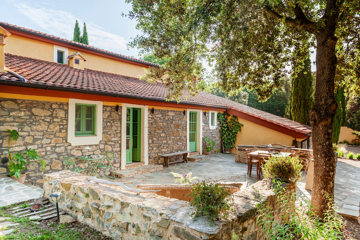 The exterior of a charming stone building is displayed with a terracotta roof and green window frames. A shaded patio area includes seating arrangements, surrounded by vibrant greenery. Natural stone walls add rustic appeal, while the setting invites relaxation and connection with nature.