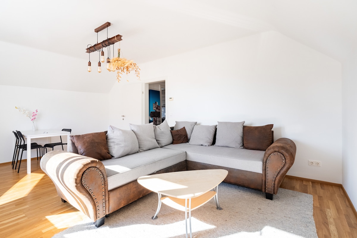 A spacious living area features a large, L-shaped sofa with several gray cushions. A minimalist round coffee table sits in front of the sofa on a textured area rug, with light streaming in from the bright, open space accented by a simple pendant light.