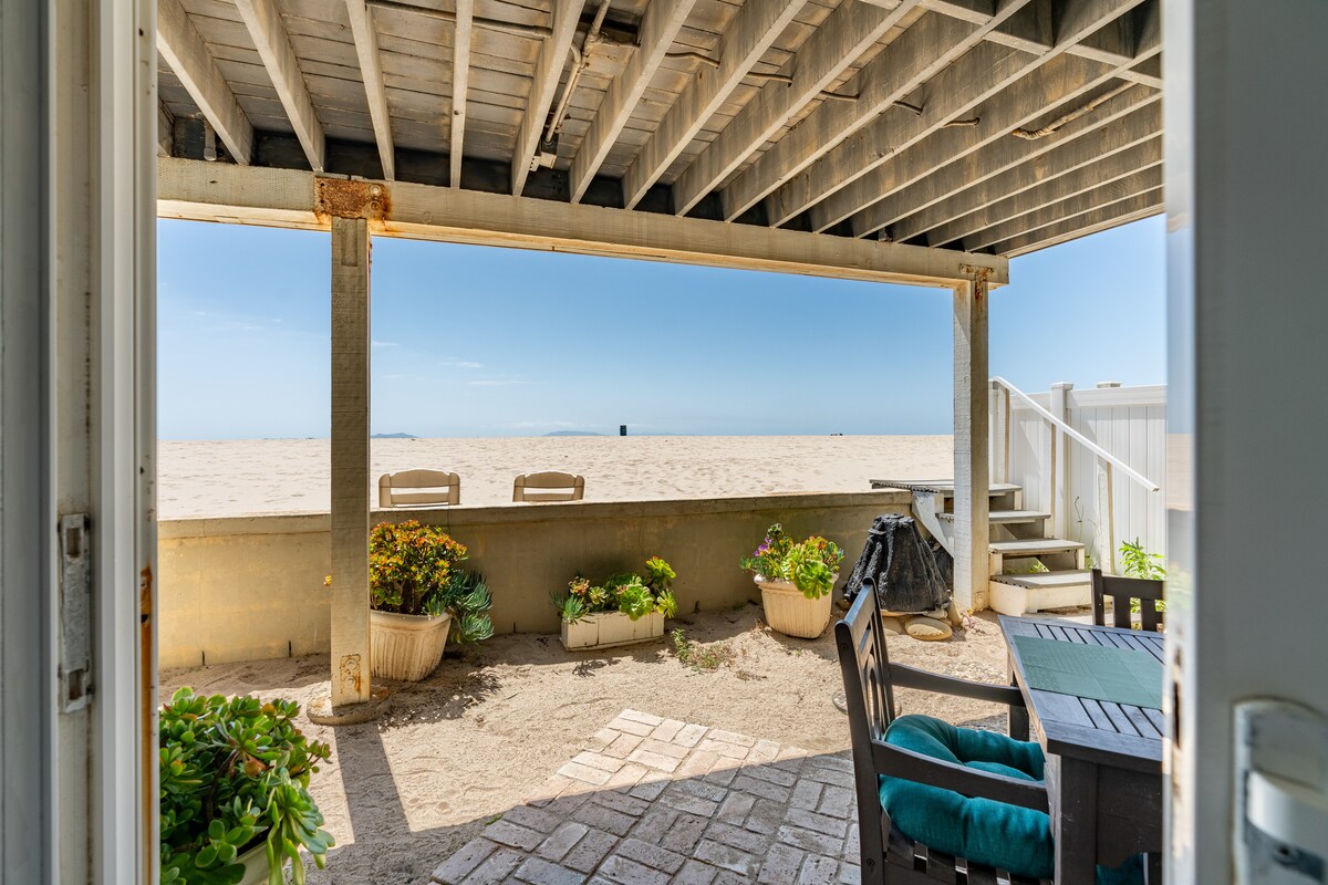 An outdoor seating area features a wooden table and chairs set against a backdrop of sandy beach and ocean views. Planters with green plants are placed nearby, providing a touch of nature. The scene is framed by the entryway, emphasizing an open and inviting atmosphere.