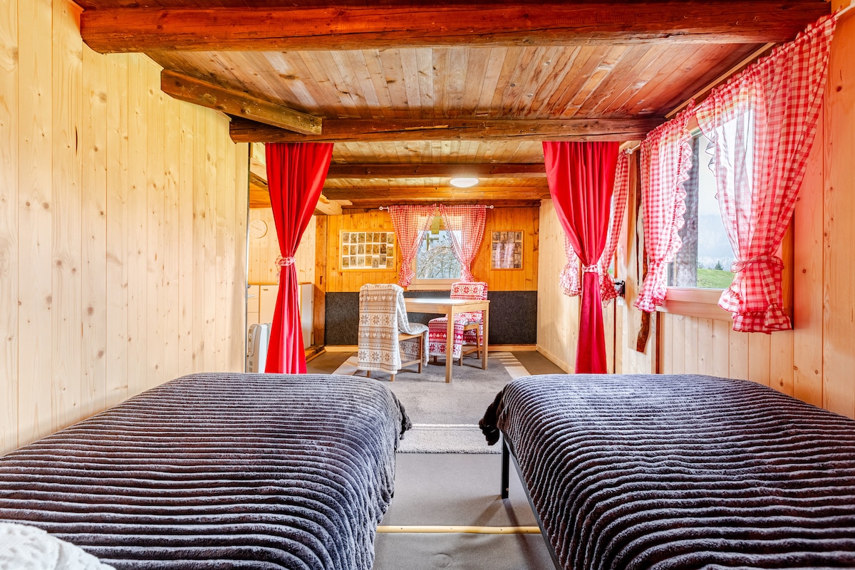 The sleeping area features two single beds with dark bedding, separated by wooden beams. Soft curtains in a vibrant red frame the windows, allowing natural light to illuminate the wood-paneled walls. A cozy seating area is visible in the background, enhancing the warm atmosphere.