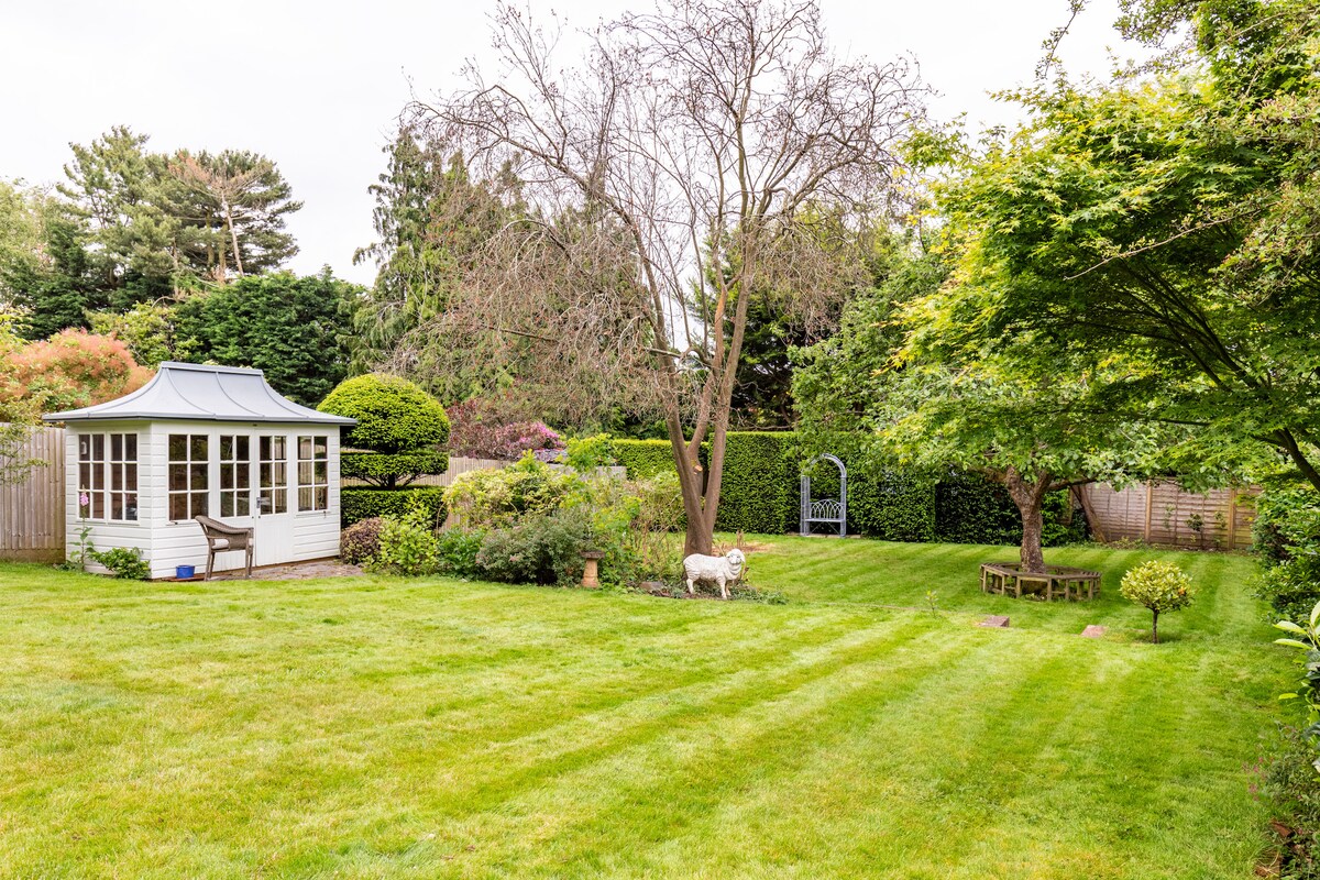 A spacious garden area features well-manicured grass, lined with tree plantings and vibrant shrubs. A white wooden gazebo is positioned to one side, complemented by a simple seating area nestled among the greenery. A path meanders through the garden, creating an inviting outdoor space.