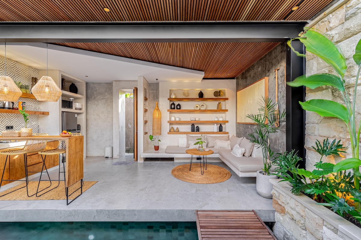 A modern living space features an open layout with sleek design elements. A comfortable seating area is adorned with natural tones, complemented by decorative shelves displaying various items. Bright greenery surrounds the space, enhancing the serene ambiance. A kitchen area is visible in the background.
