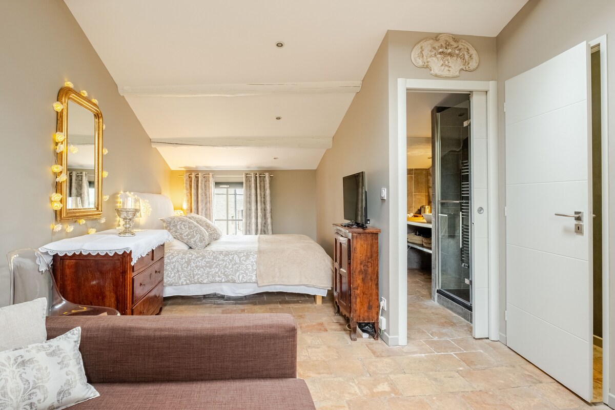 A spacious room is presented featuring a large bed dressed in neutral tones, complemented by soft bedding. A wooden dresser and a mirror enrich the decor. Natural light enters through large windows, illuminating a cozy seating area and an ensuite bathroom visible through an open doorway.