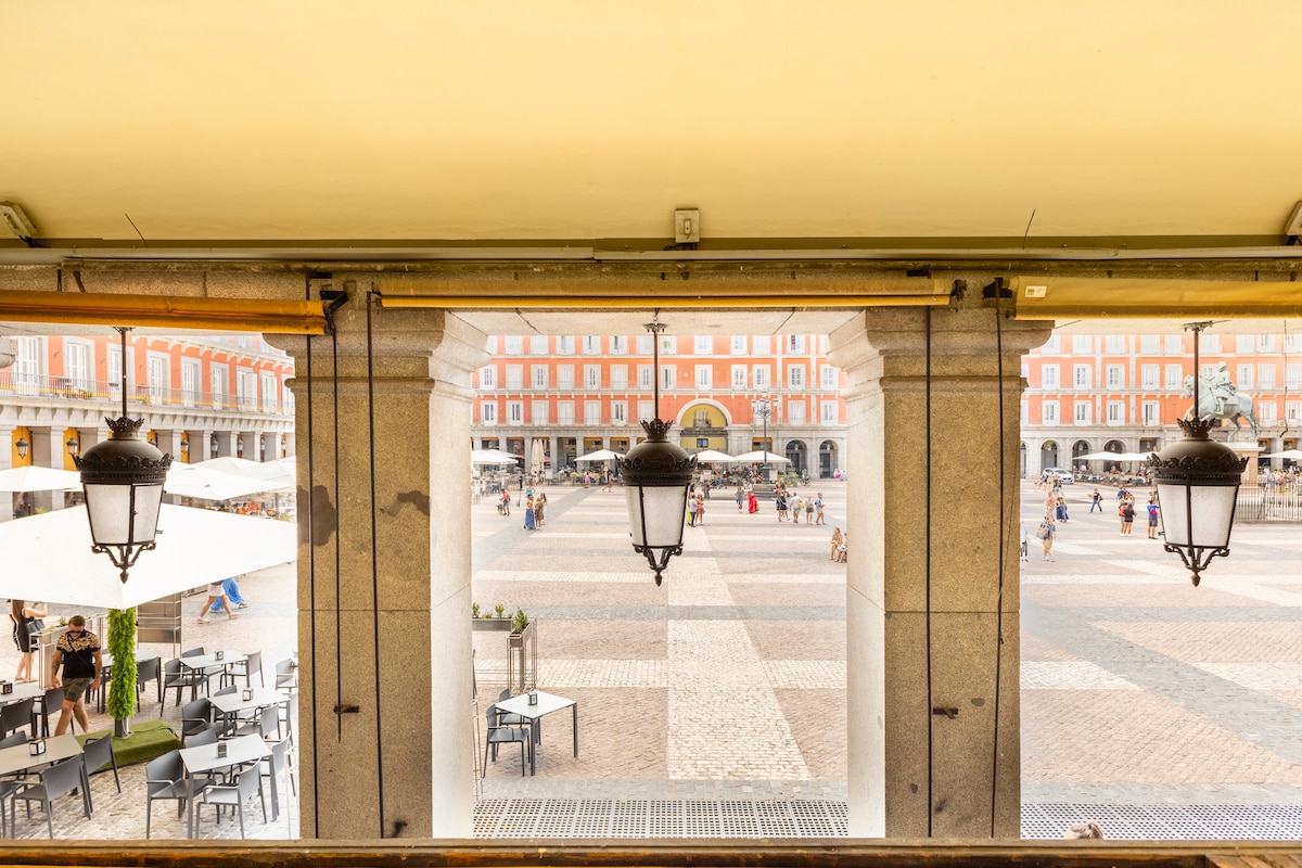 Views of Plaza Mayor are framed by two elegant lanterns hanging from arched columns. The spacious square features a cobblestone design with scattered seating arrangements, while visitors can be seen strolling at a leisurely pace. The vibrant buildings of the plaza provide a charming backdrop.