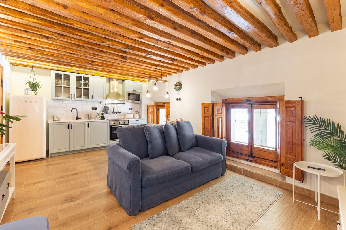 A spacious living area features exposed wooden beams overhead, contributing to a rustic charm. A comfortable blue sofa is positioned near a window with wooden shutters. Light-colored cabinetry and appliances are visible in the background, alongside a small rug that adds a touch of softness.