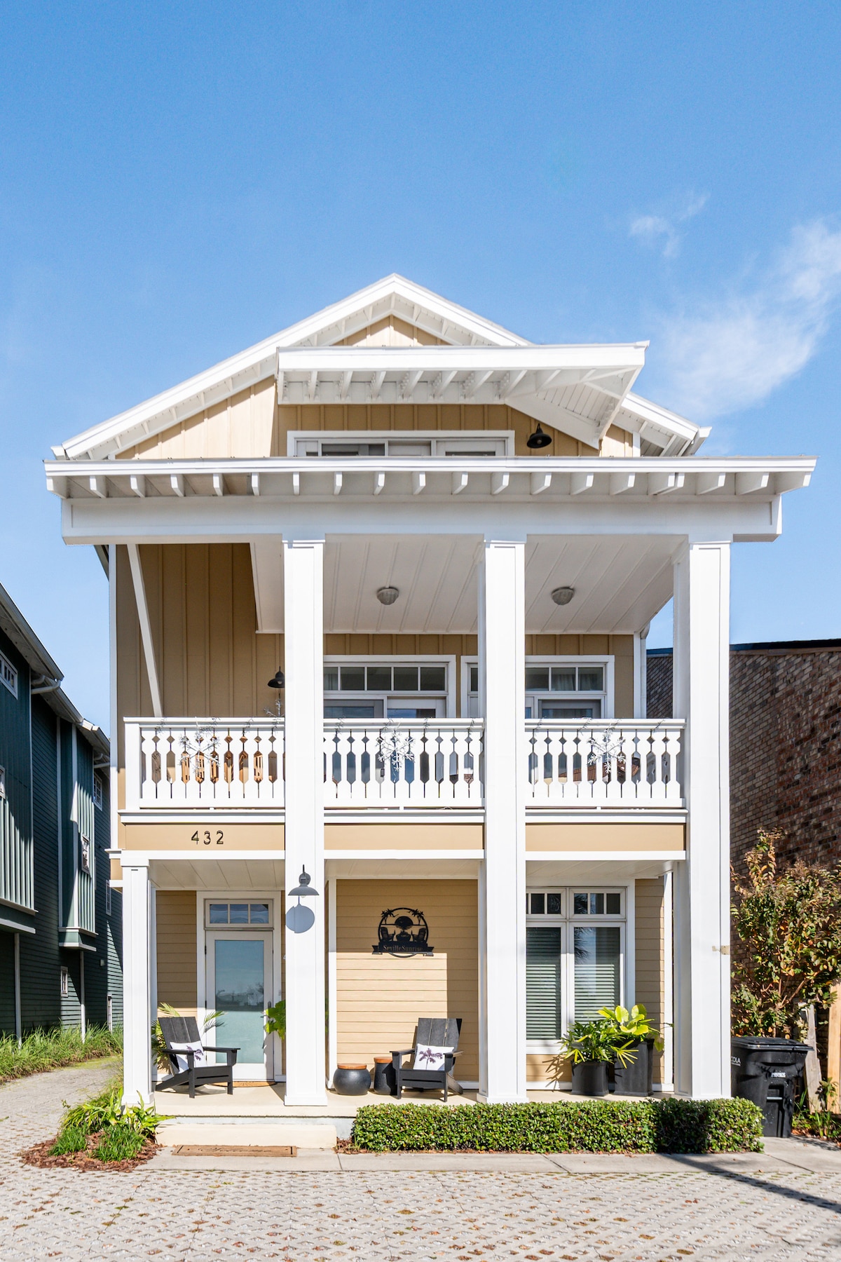 The charming exterior of a two-story villa presents a light-colored facade with white accents. A covered balcony showcases elegant railings, while two chairs are placed on the ground level porch. Lush greenery surrounds the entrance, offering a welcoming atmosphere.