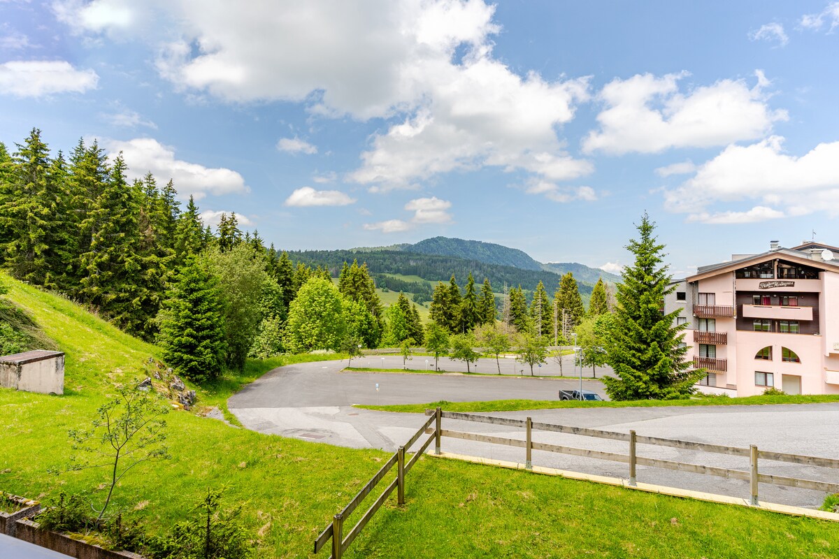 A panoramic view captures a serene landscape featuring rolling green hills and a treeline in the distance. A multi-story building is visible on the right, with a parking area seen below. The sky is dotted with fluffy clouds, enhancing the peaceful setting.
