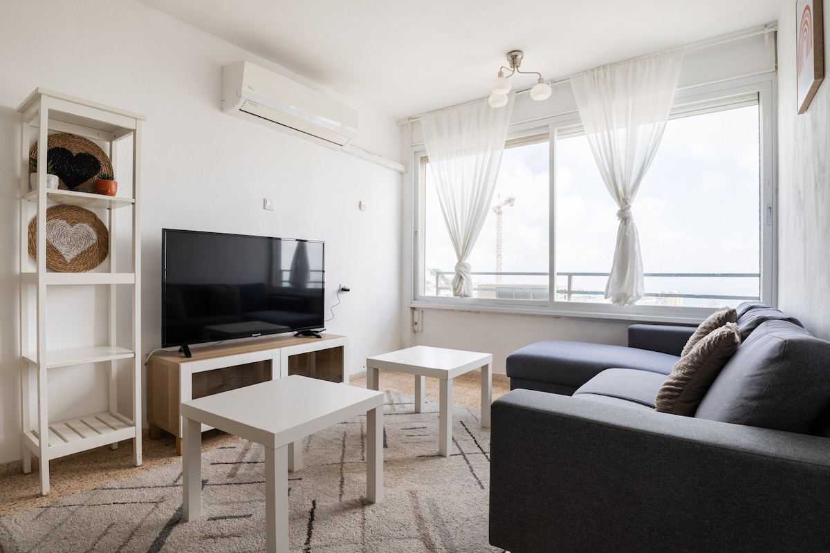 A bright living area features large windows that offer views of the outdoors. A flat-screen television is positioned on a modern stand. Two coffee tables are placed in front of a comfortable sectional sofa, complemented by a cozy armchair and decorative shelving.
