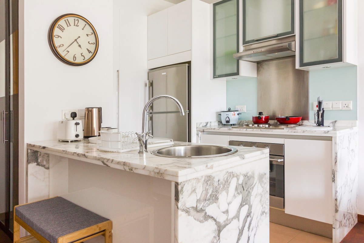 A modern kitchen space is highlighted by sleek marble countertops and contemporary cabinetry. The stainless steel refrigerator and multiple cooking appliances are visible, along with a double sink and a small wooden stool for seating. A clock adorns the wall, providing a functional accent.