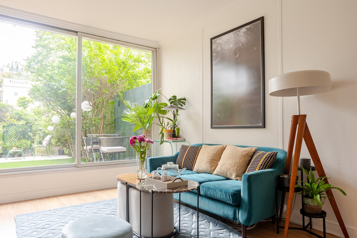 A spacious living area is presented with a large window overlooking a private garden. A teal sofa is paired with a round coffee table, and a stylish lamp stands nearby. A vase of fresh flowers adds a touch of color to the serene setting.