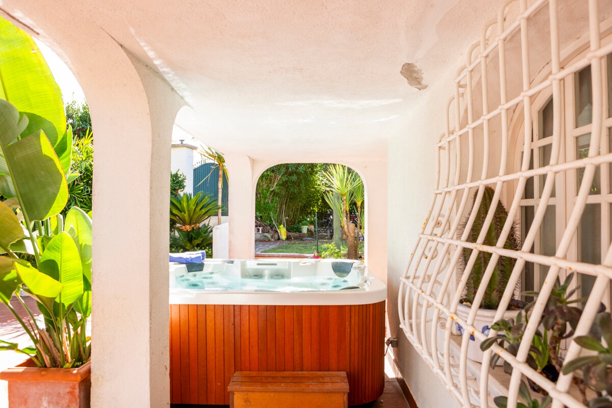 A shaded outdoor area features a wooden hot tub surrounded by lush plants. The space is framed by an arched entrance and a decorative window grating, allowing natural light to flow through. The inviting tub reflects the surrounding greenery in its water.