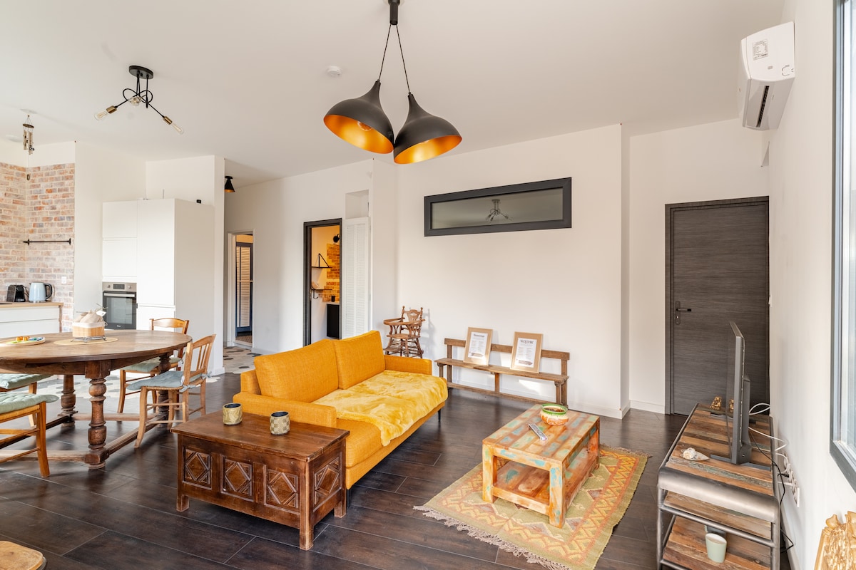 A spacious living area is seen with a large orange sofa facing a wall-mounted television. A wooden table and chairs are situated nearby, with light streaming in from a window. The open layout features stylish lighting fixtures and a modern kitchen in the background.