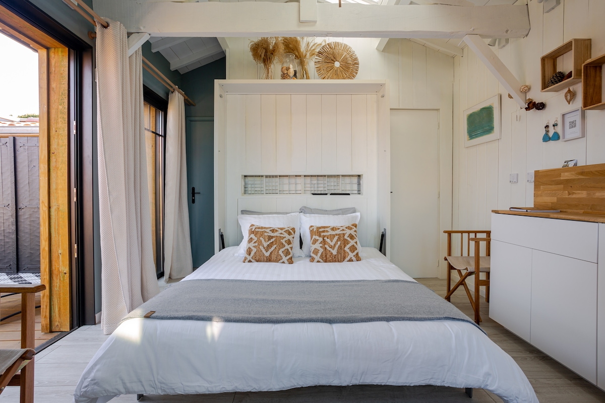 A bright and airy space is depicted, featuring a spacious bed adorned with decorative pillows. The soft tones of the walls are complemented by natural elements in the decor. A small kitchen area is visible, along with a wooden chair and sliding glass doors that lead to an outdoor terrace.