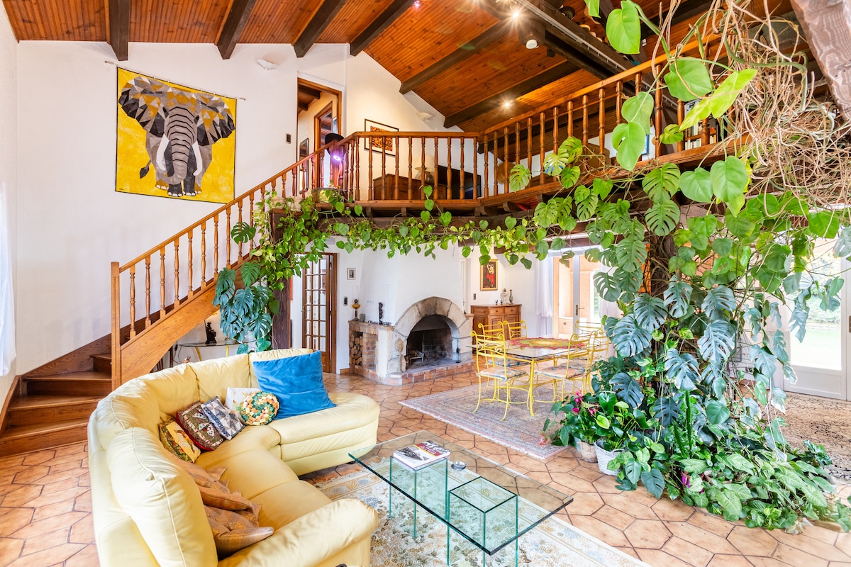 A spacious living area features a yellow sofa, glass coffee table, and lush indoor plants. A large staircase leads to a mezzanine level. Artistic decor, including an elephant painting, enhances the room's character. Natural light floods through large windows, creating a bright environment.