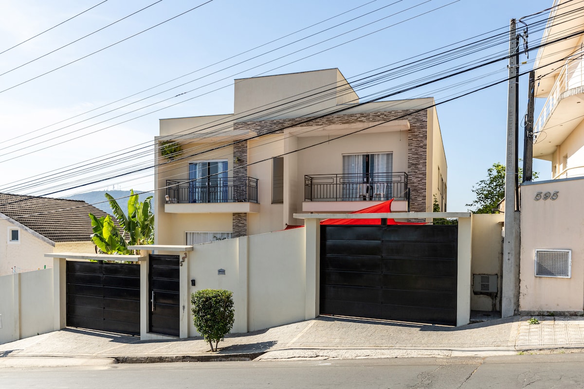 A modern two-story home is showcased, featuring light-colored walls and decorative stone accents. The building is set back from the street, with large black gates marking the entrance. A vibrant red awning provides shade over the balcony. Lush greenery is present in the front yard.