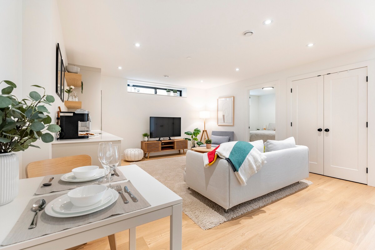 A modern living space showcases a cozy sofa and lounge chair, positioned in front of a smart TV. A dining area features a table set for two with dinnerware. Light-colored walls and warm wooden flooring enhance the bright and clean atmosphere.