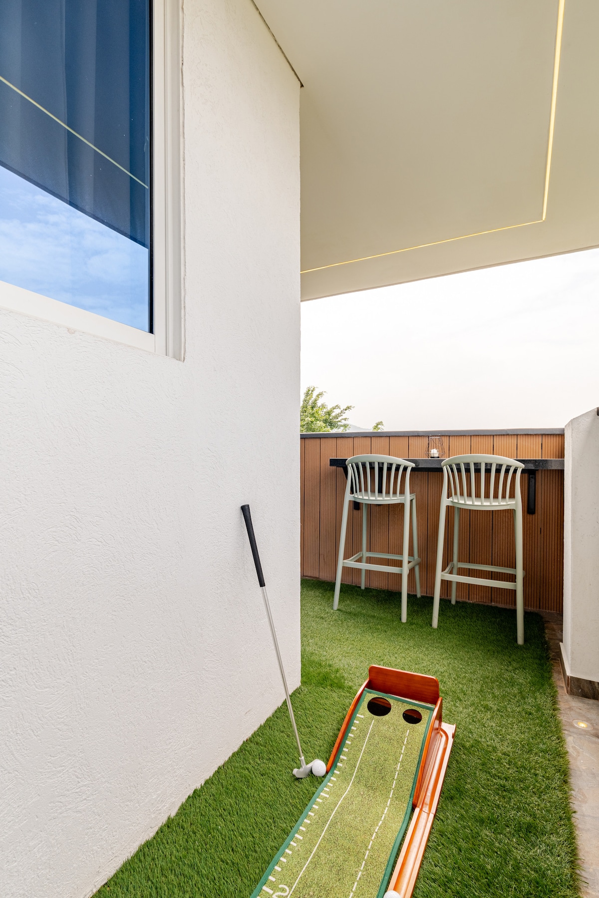 A small balcony area features artificial grass, a putting green, and a golf club placed next to a white bar height table with two chairs. The wall behind is painted white, and a window allows natural light to fill the space.