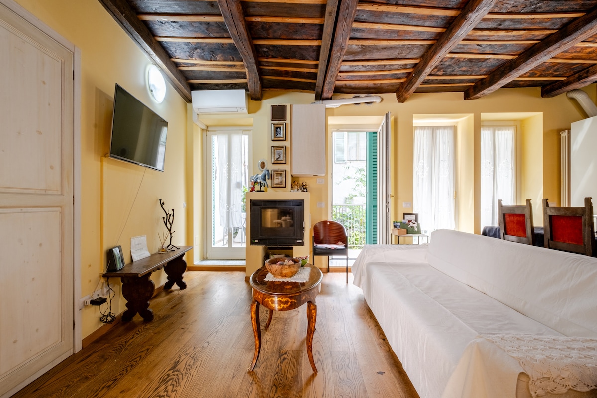An inviting living space features wooden beams overhead and a cozy fireplace. A light-colored sofa is positioned across a decorative coffee table. Large windows allow natural light to illuminate the room, with views of greenery from the courtyard visible through the open doors.