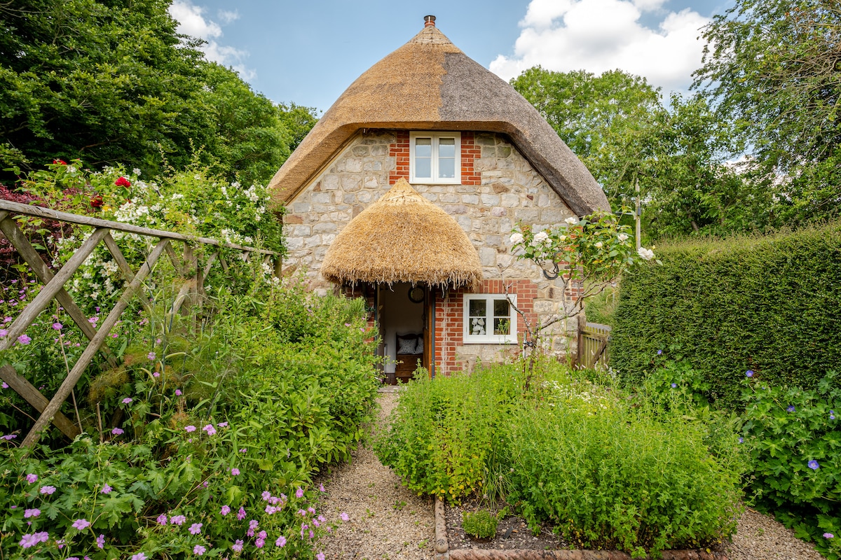 The charming cottage features a thatched roof and rustic stone walls, surrounded by vibrant greenery and colorful flowers. A gravel path leads to the welcoming entrance, framed by lush plants and a wooden fence, creating a serene and inviting atmosphere.