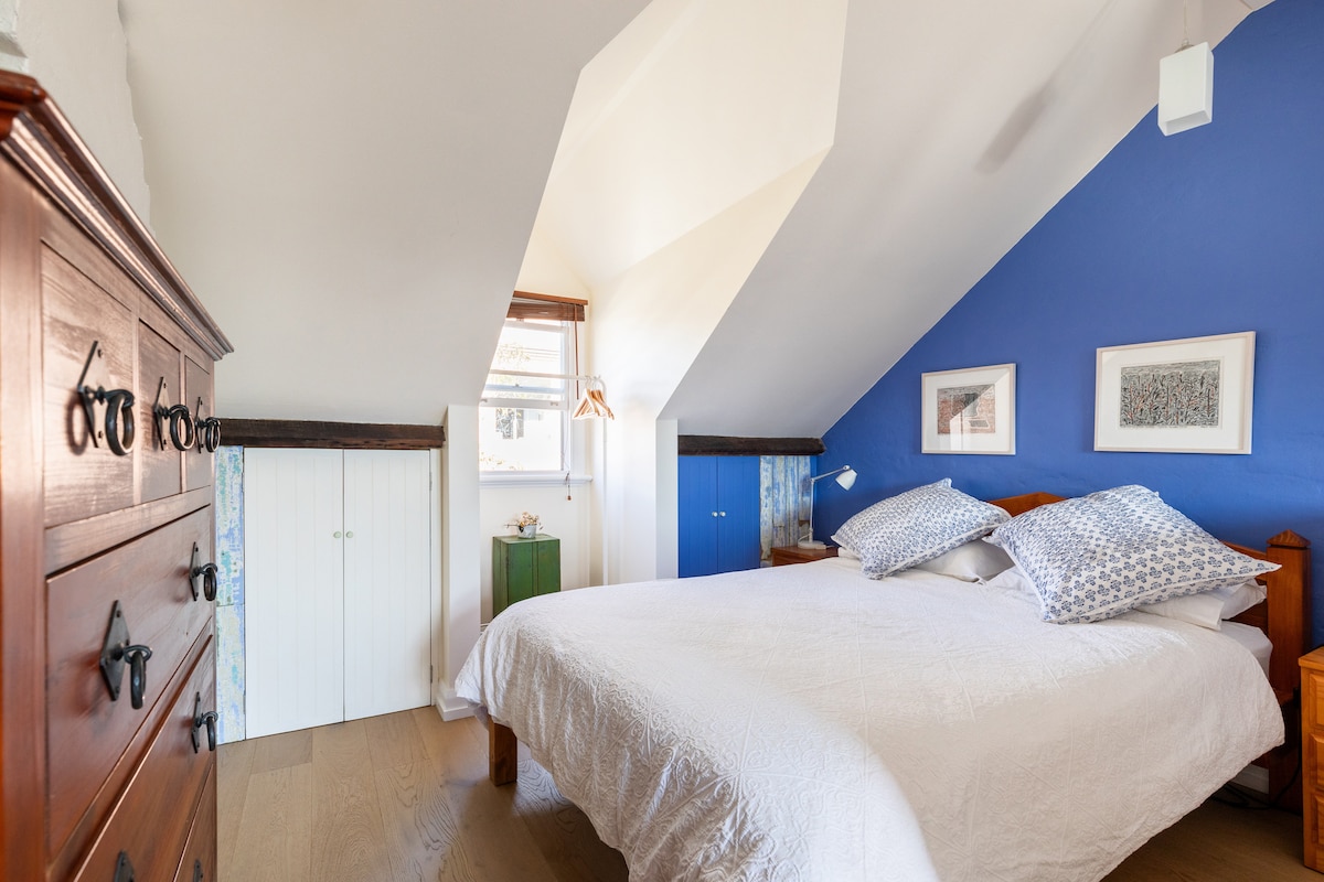An attic bedroom is displayed with sloped ceilings and vibrant blue walls. A spacious bed is centered in the room, adorned with patterned pillows. Wooden furniture, including a chest of drawers and bedside tables, is positioned alongside two framed artworks on the wall. Natural light filters through a window.
