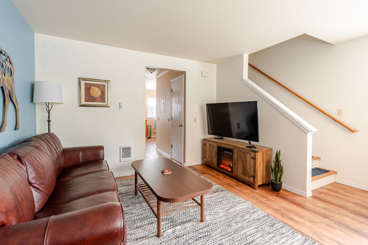 A spacious living room features a brown leather sofa and a wooden coffee table positioned in front of a large flat screen TV. Natural light fills the space, with a staircase visible in the background leading to the upstairs landing.