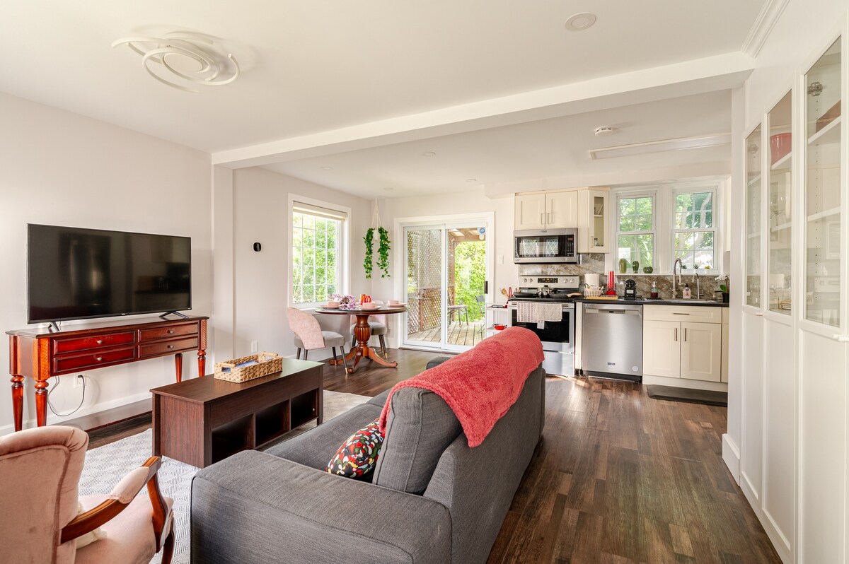 A spacious living area features a comfortable gray sofa and a wooden coffee table. Large windows allow natural light to fill the room. A dining area is visible in the background, alongside a modern kitchen equipped with stainless steel appliances.